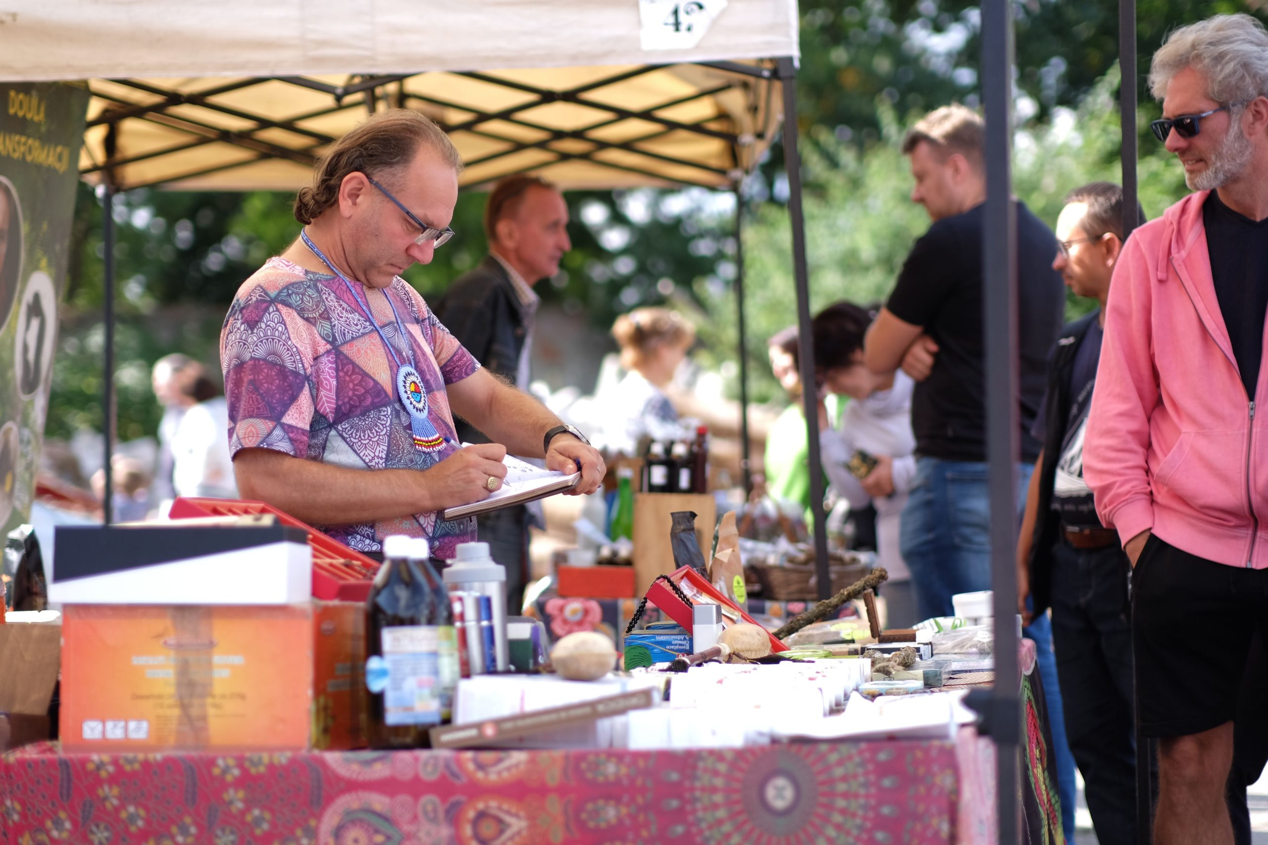 A vendor standing at a market stall with various products in an outdoor bazaar.